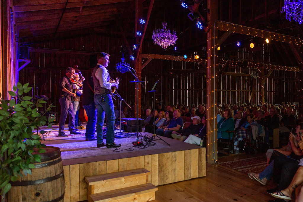 The band Balsam Range performing for The Series at Lavender Hill Farm on the stage in their events barn. This is side view of the stage and you can see purple lighting, chandeliers, twinkle lights on the barn's beams and a full audience.