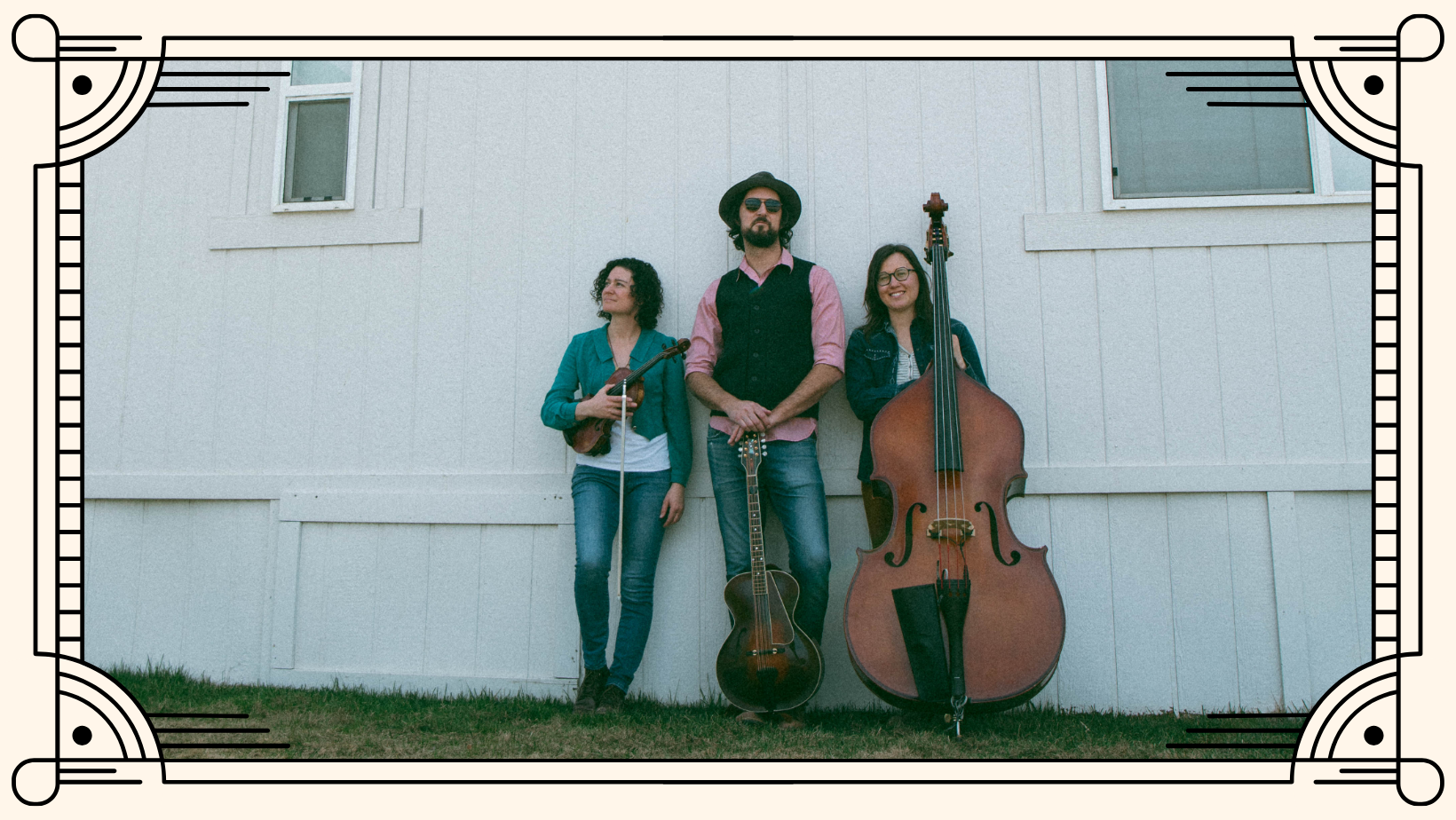 Photo of the trio band, Stillhouse Junkies standing against a white building holding instruments.